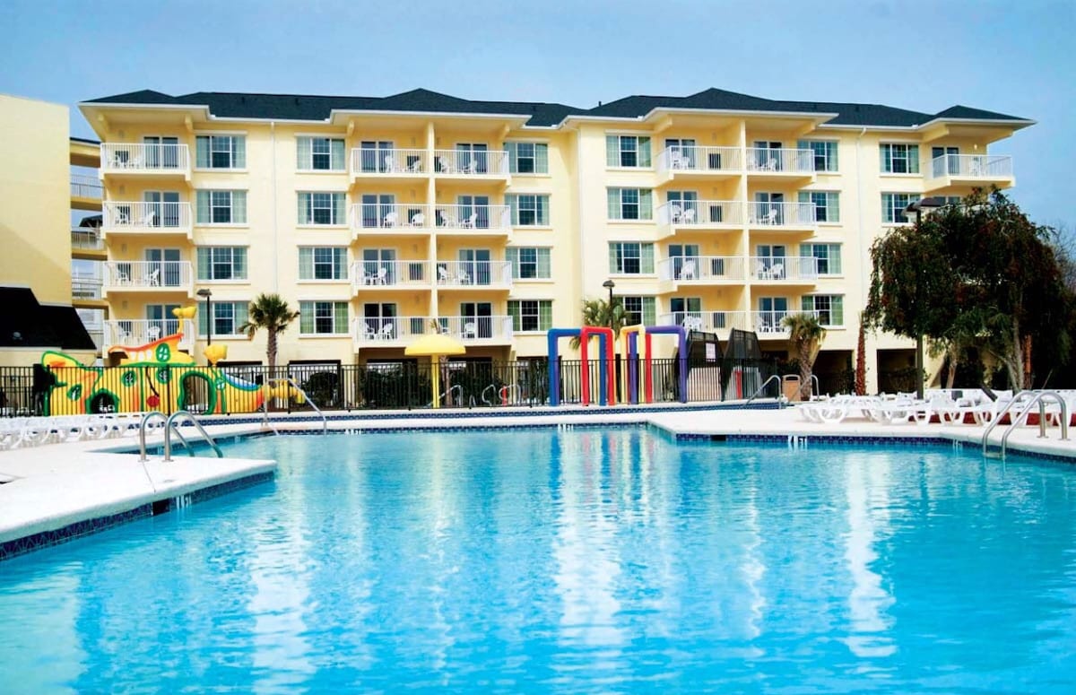 A well-maintained pool area is visible, surrounded by lounge chairs and colorful play structures for children. The multi-story building stands in the backdrop, featuring multiple balconies and large windows, reflecting a sunny day. Palm trees are positioned around the pool, adding to the resort-like atmosphere.