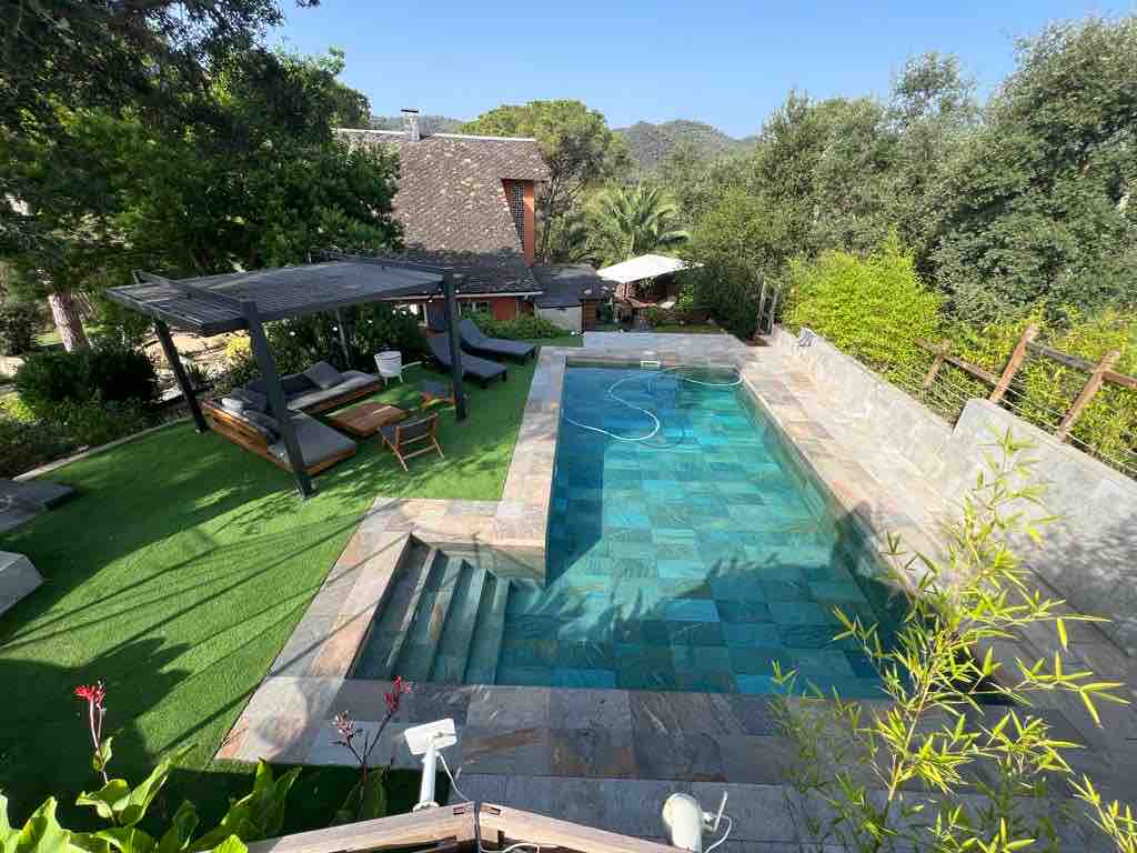 The outdoor area features a private pool surrounded by landscaped greenery. Comfortable lounge chairs are positioned under a shaded structure nearby. The pool’s turquoise water reflects the surrounding natural beauty, while steps lead down into the shallow end, creating an inviting space for relaxation.