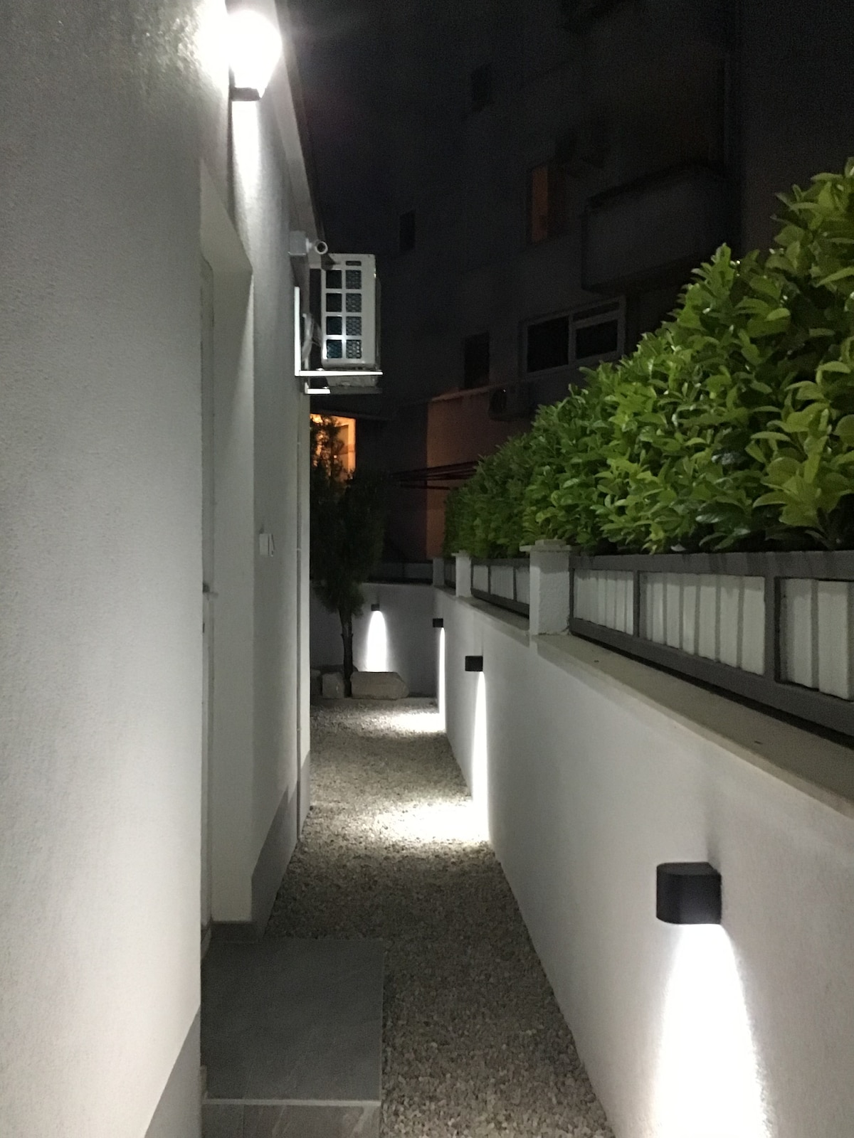 A well-lit pathway leads alongside a smooth, white exterior wall, bordered by lush greenery. Soft lighting illuminates the walkway, creating a serene atmosphere. The entrance to the guesthouse is visible at the end of the path, framed by elegant plantings.