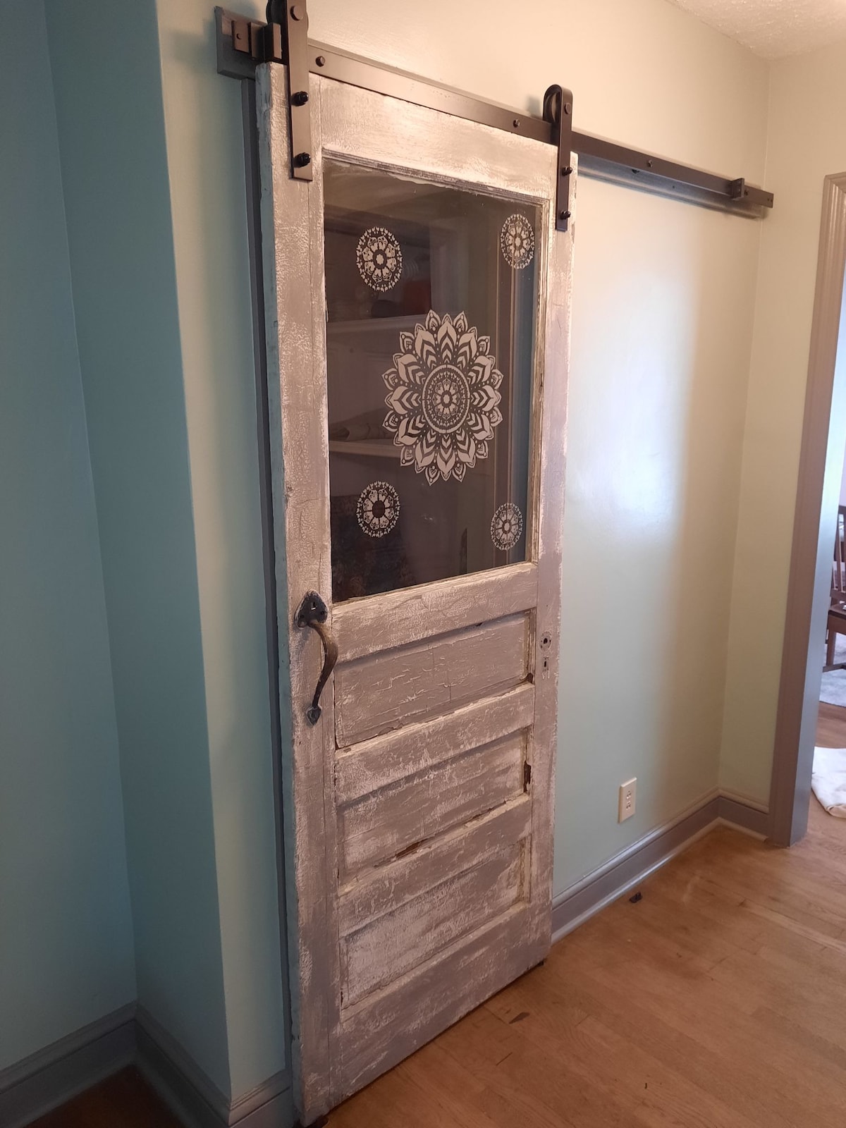 A sliding barn door with a weathered white finish is showcased, featuring a decorative central glass panel adorned with intricate floral designs. The door is set against a soft blue wall, and the wooden floor adds a rustic touch to the cottage's interior.