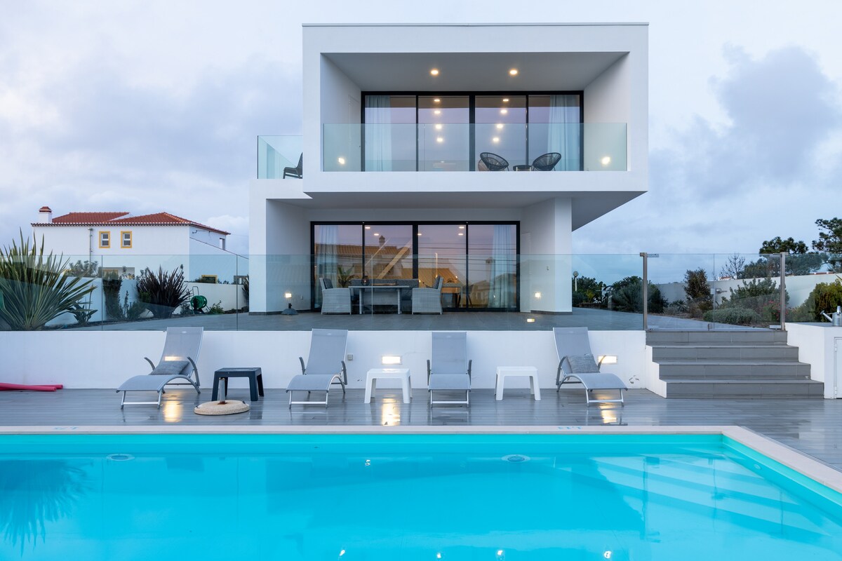 A modern structure features large glass windows on the upper level, offering outdoor seating on a balcony. Surrounding the inviting pool, multiple lounge chairs are arranged, with a mix of colors and designs. Lush greenery adds a natural element to the contemporary landscape.