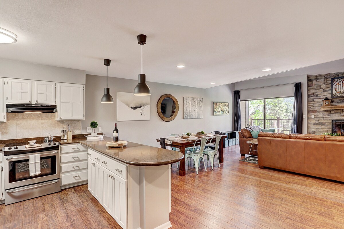 The open-concept living area features a modern kitchen with white cabinetry and dark countertops. A dining table with metal chairs is set nearby. Natural light streams in through large windows, illuminating the comfortable seating area next to a stone fireplace.