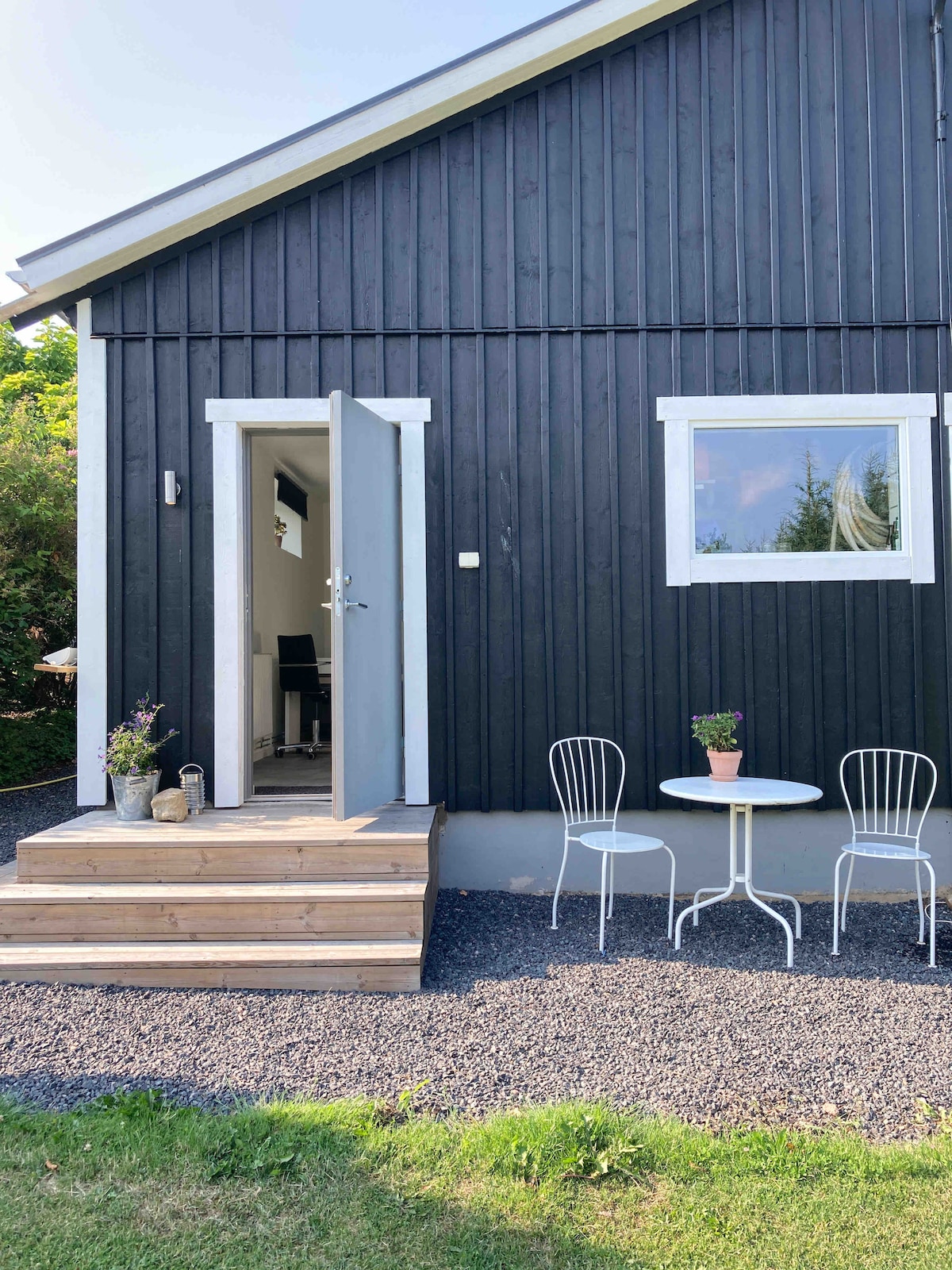 The exterior of the apartment is shown, featuring a modern wooden facade painted in dark tones. A simple wooden stairway leads up to an open door. A small round table with two chairs is positioned on the gravel surface, adorned with potted flowers.