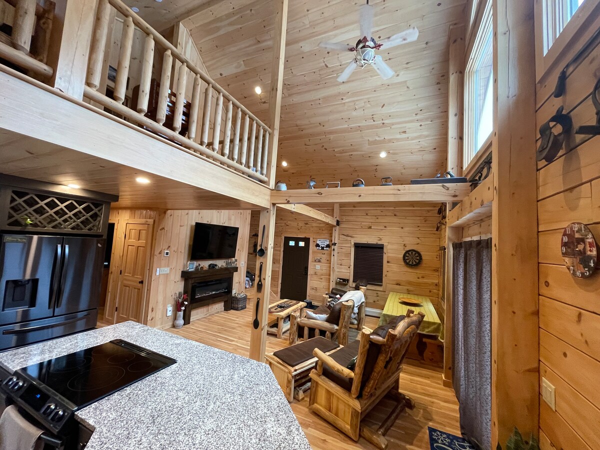 A spacious, open-concept living area showcases natural wood paneling and high ceilings. The kitchen features a granite countertop with a modern stovetop, while various seating options, including rustic wooden chairs and a couch, are visible. Large windows allow ample natural light.
