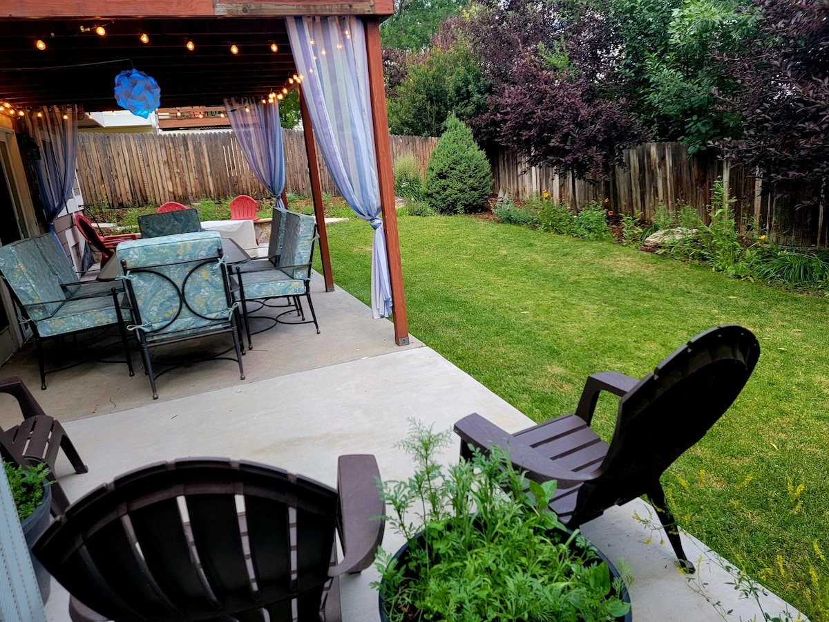 An outdoor patio area is shown, featuring a table with seating for six, surrounded by greenery. Comfortable lounge chairs are positioned nearby, inviting relaxation. Soft lighting hangs above, enhancing the space. The well-maintained lawn showcases a variety of plants and trees at the back.