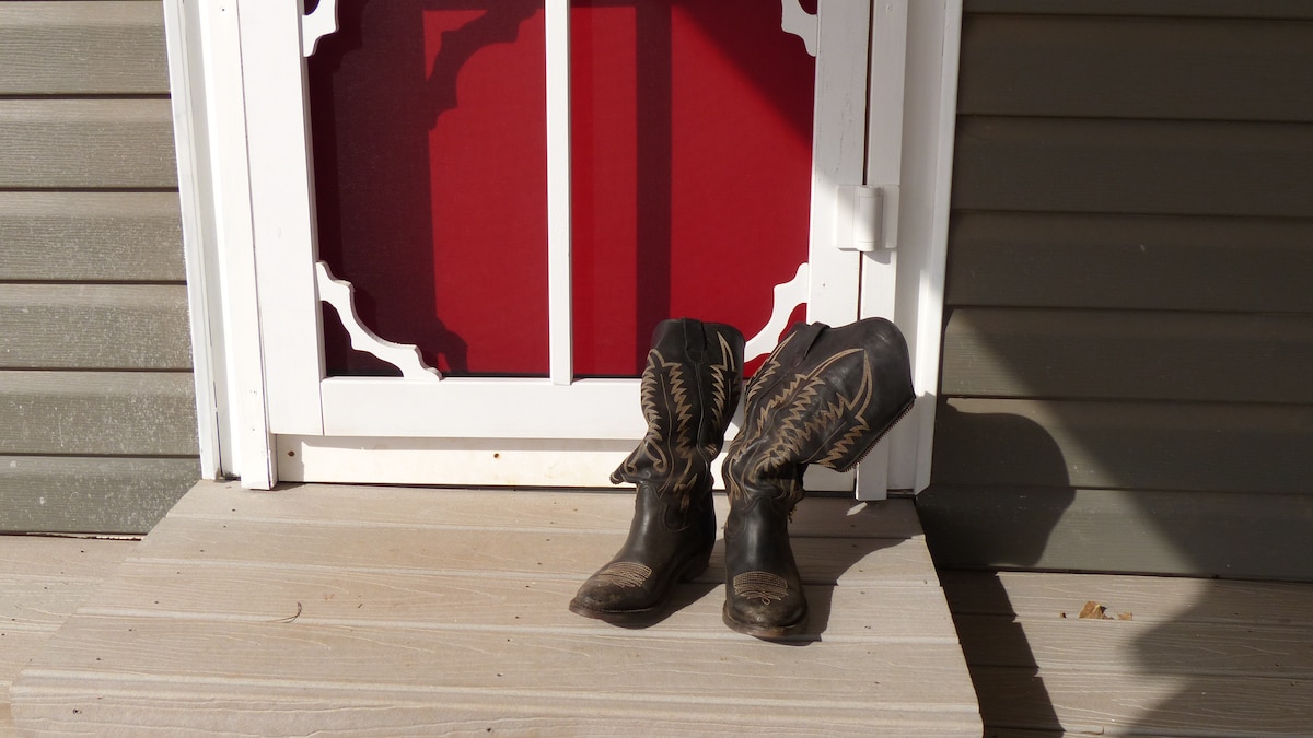 A pair of classic cowboy boots is placed on the wooden porch in front of a red door. The boots, showing signs of wear, add a rustic charm to the entrance, which is framed by decorative white trim.