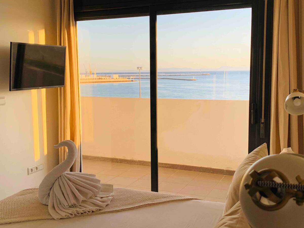 A serene view of the sea is visible through a large glass door, leading to a private terrace. The room features a spacious bed with crisp linens, and a television mounted on the wall, creating a calm and comfortable atmosphere.