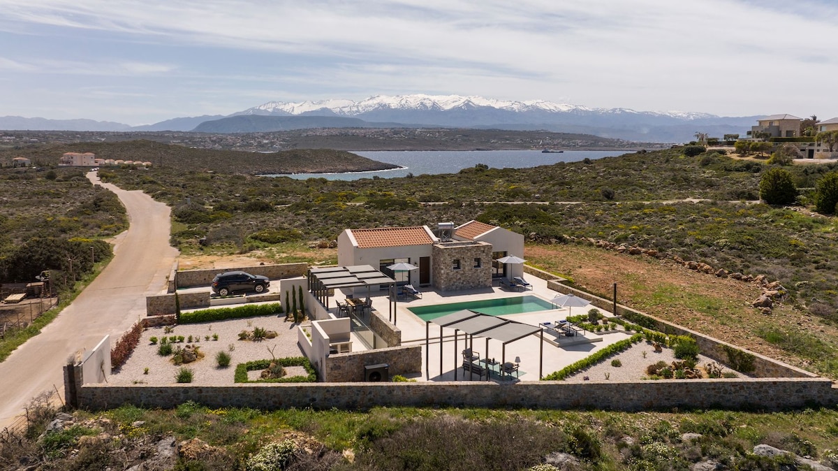 The exterior view captures a modern villa surrounded by landscaped grounds, featuring a spacious outdoor area with a swimming pool and lounge seating. Majestic mountains are visible in the background, along with a serene body of water, creating a tranquil backdrop.