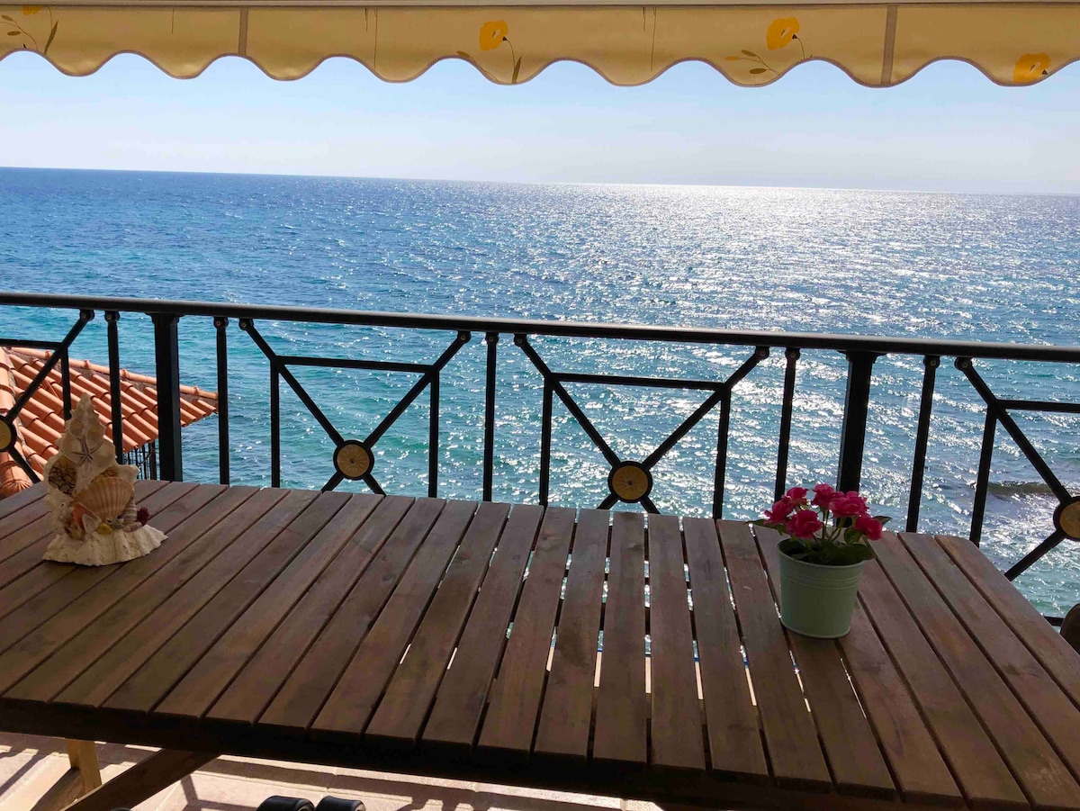 A wooden table is positioned on a balcony overlooking the shimmering Aegean Sea under a bright blue sky. A potted flower sits on the table, adding a natural touch. The scene is framed by a canopy that provides shade, enhancing the outdoor dining experience.
