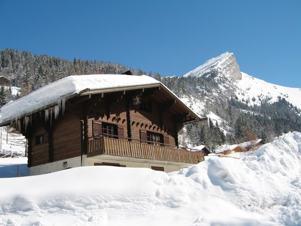 The wooden chalet is surrounded by a snowy landscape, with a sloping roof adorned with icicles. A spacious balcony offers views of the mountain backdrop, while tall pine trees complete the serene alpine setting. The clear blue sky enhances the peaceful atmosphere of the location.