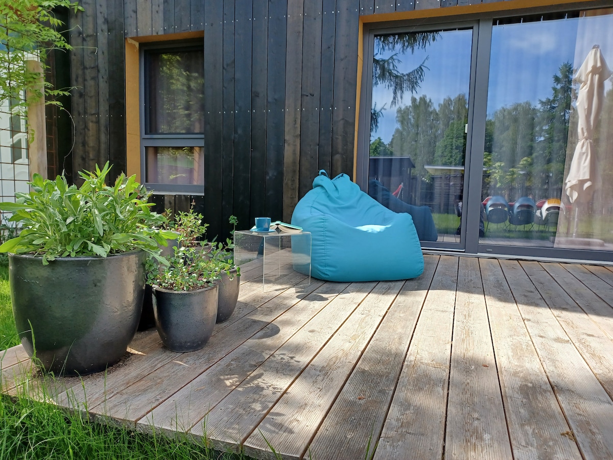 A wooden deck is shown, featuring a vibrant blue bean bag chair and potted plants. Large glass doors provide a view of the green garden area beyond. Sunlight streams gently onto the deck, creating a peaceful outdoor space.