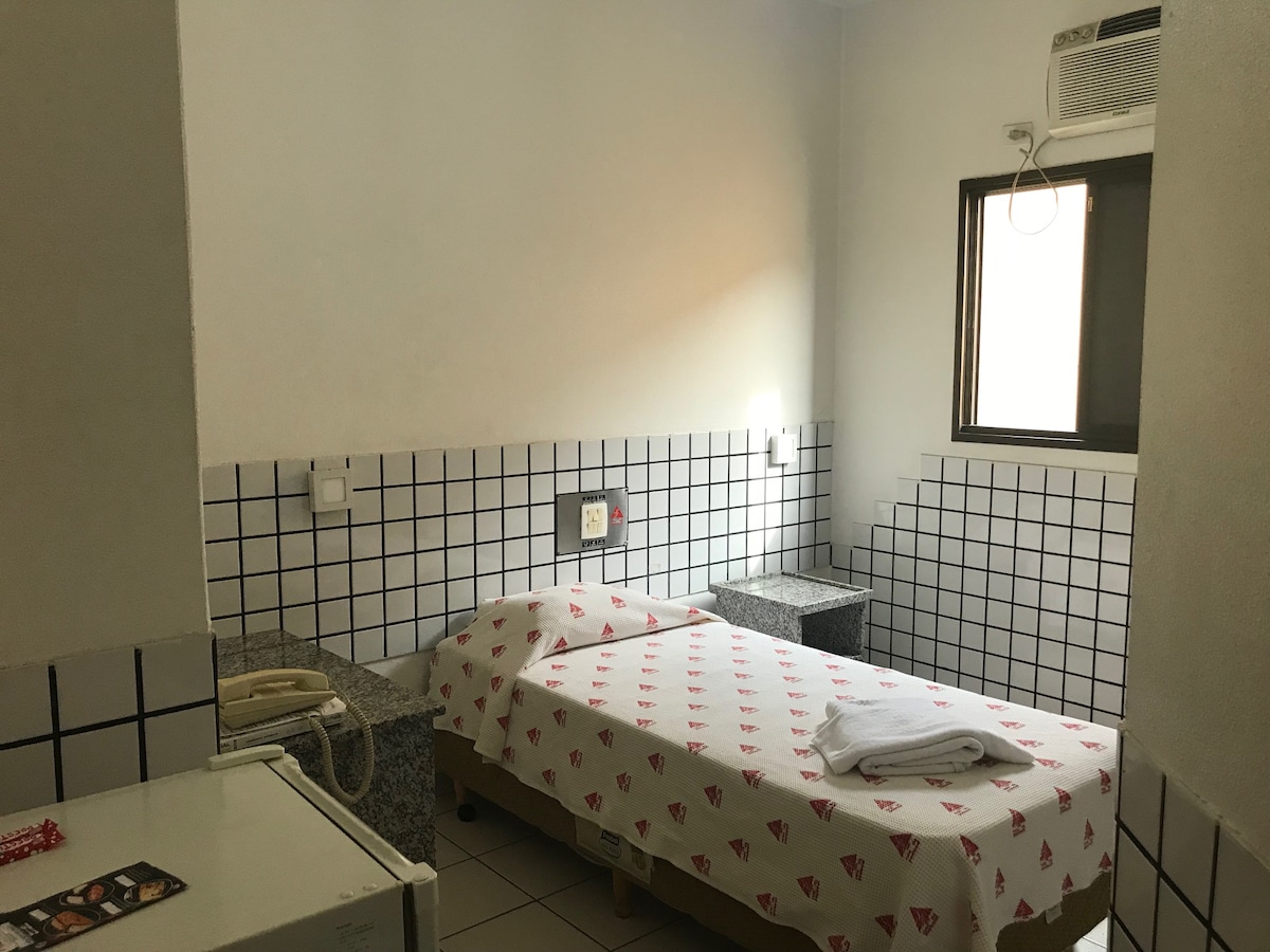 A simple sleeping area features a bed covered with a white sheet adorned with a red pattern. Two granite-topped nightstands stand on either side of the bed, and a window allows natural light to enter the room.