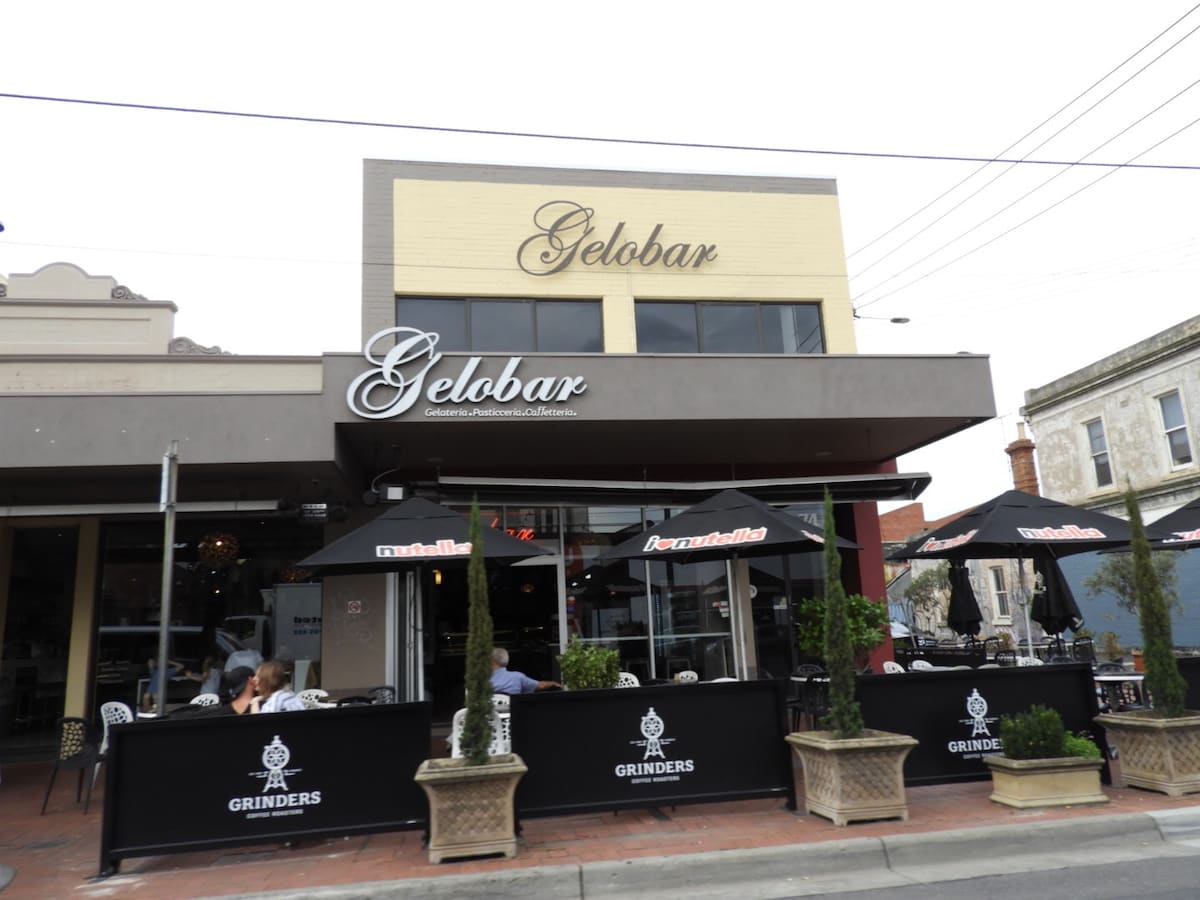 The exterior of Gelobar showcases a two-level building with a warm yellow façade. Outdoor seating is available under black umbrellas marked with the Grinders logo, arranged around potted plants. Patrons can be seen enjoying their meals at the sidewalk café, creating a lively atmosphere.