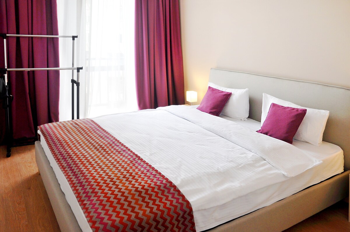 A spacious bed is dressed in crisp linens, complemented by decorative pillows in shades of purple. Light filters in through the sheer curtains, illuminating the room with a warm glow. A clothing rack is positioned in the corner, enhancing the room's functionality.