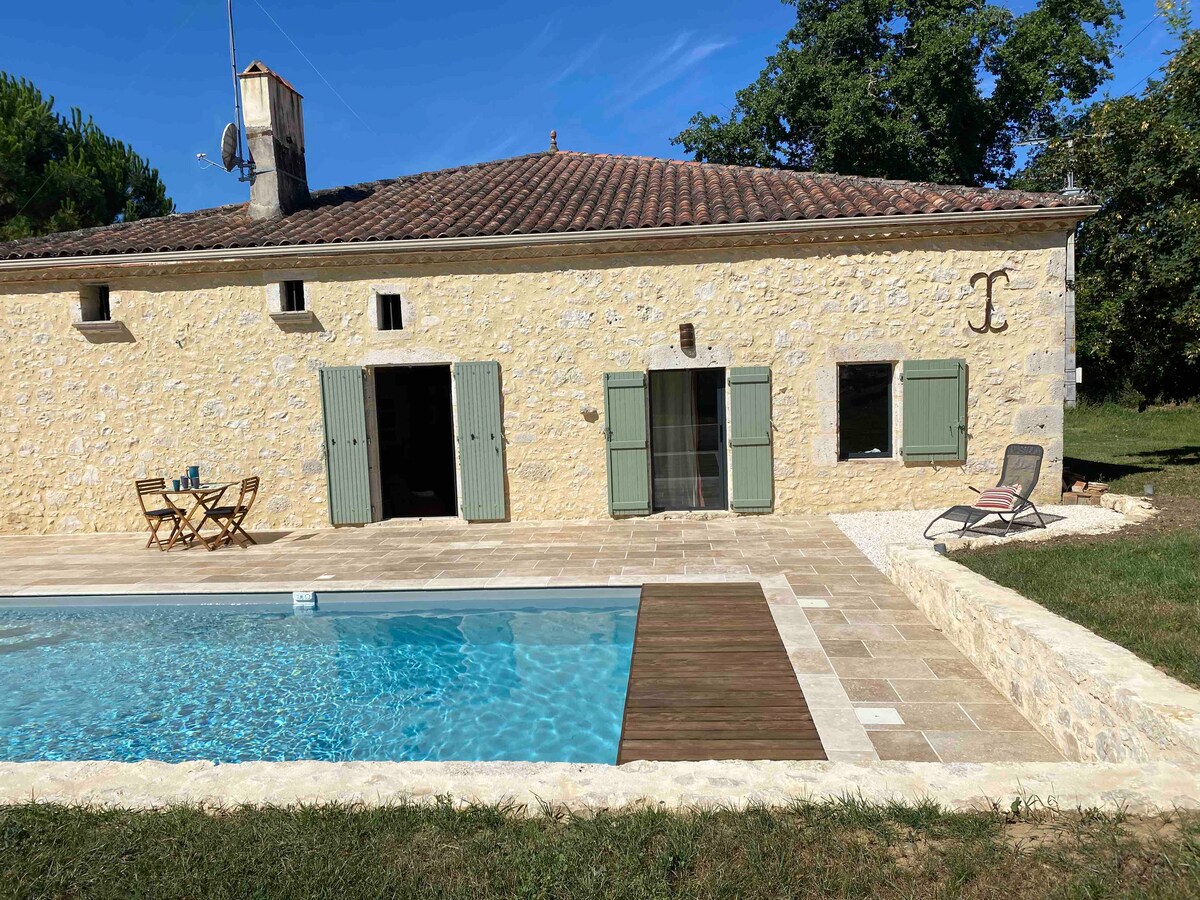 The exterior of a stone villa is featured, showcasing light yellow walls and green shutters. A swimming pool is positioned in front, surrounded by a tiled patio and a wooden deck. Two outdoor chairs and a table are visible nearby, set in a grassy area.
