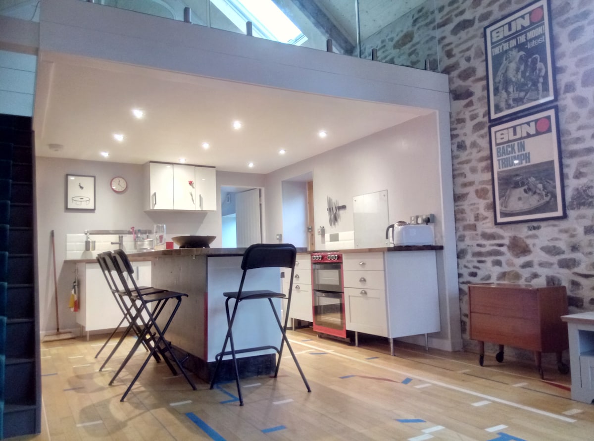 An open-plan kitchen area features a generous island with seating for two black chairs. Modern appliances, including a stove and kettle, are arranged along the white cabinetry. Exposed stone walls add character, while bright overhead lights enhance the spacious feel. A large skylight allows natural light to fill the space.