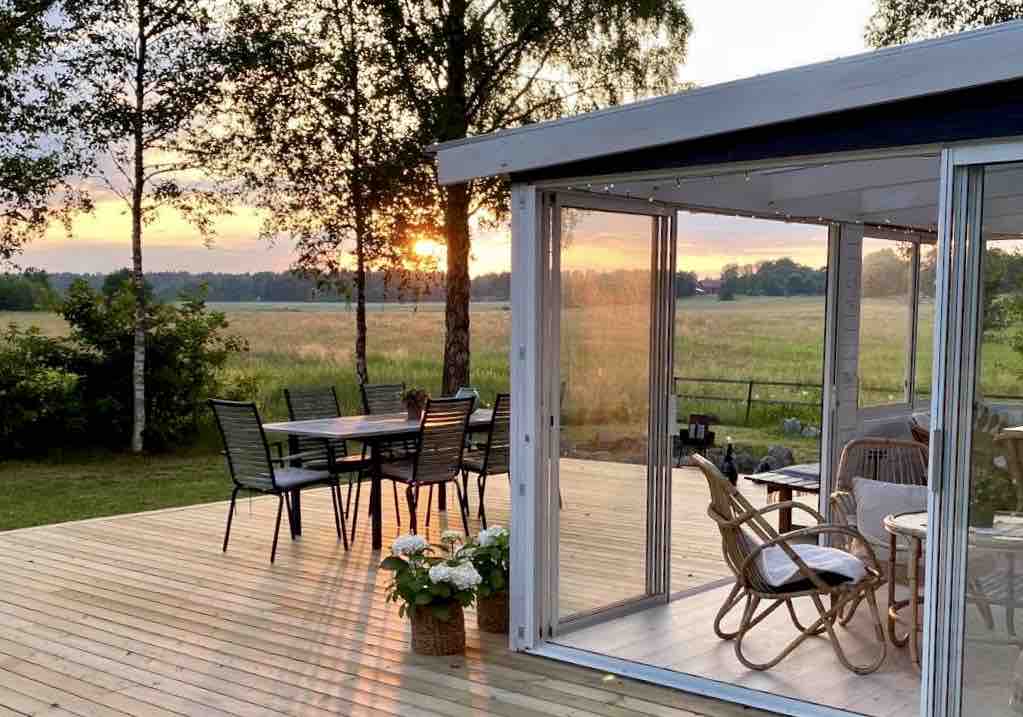 A glass-enclosed patio offers views of a sunset over a grassy field. The space includes a dining table with six chairs and a seating area featuring a circular chair. Potted plants are arranged neatly on the wooden deck, enhancing the connection to nature.
