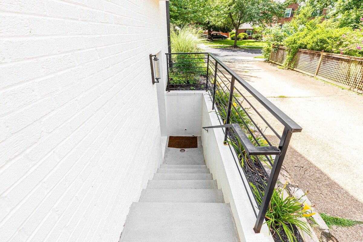 A concrete staircase is visible, leading down to the lower level apartment. A metal railing lines the steps for support. Lush greenery and landscaping are present along the path, while a paved driveway is seen at the top of the stairs.