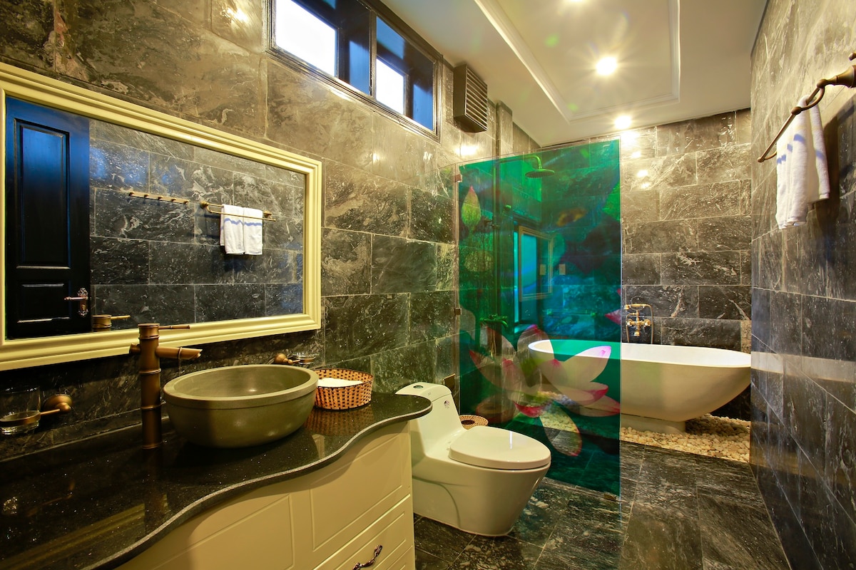 A modern bathroom featuring dark polished marble walls and floors. A spacious bathtub sits beside a glass partition adorned with floral designs. A countertop with a unique sink and basin is placed next to a contemporary toilet. Natural light filters through a window above.