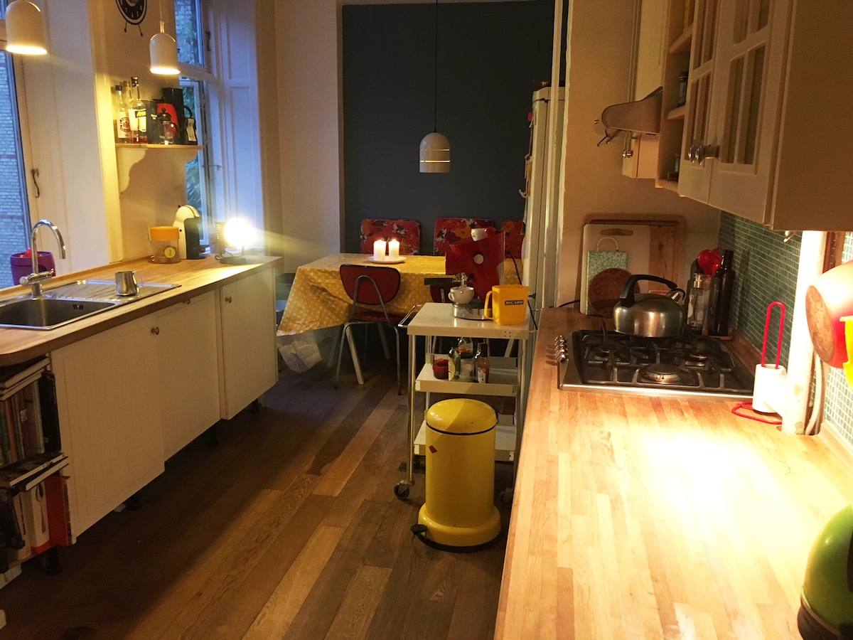 A well-lit kitchen features light wood flooring and ample counter space. A gas stove and a sink are visible, with various kitchen utensils arranged nearby. A dining area with a patterned tablecloth and colorful chairs is situated in the background, creating an inviting space for meals.