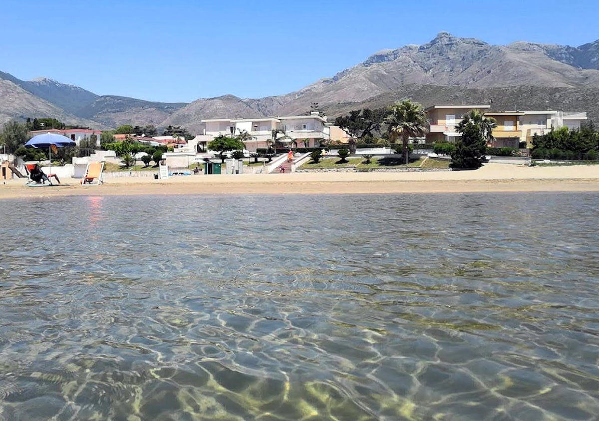 The image captures a serene beach scene with clear waters, reflecting the sandy shore and a backdrop of distant mountains. Colorful umbrellas and lounge chairs are positioned along the beach, providing spaces for relaxation. The shoreline is framed by well-maintained greenery and beachfront residences.