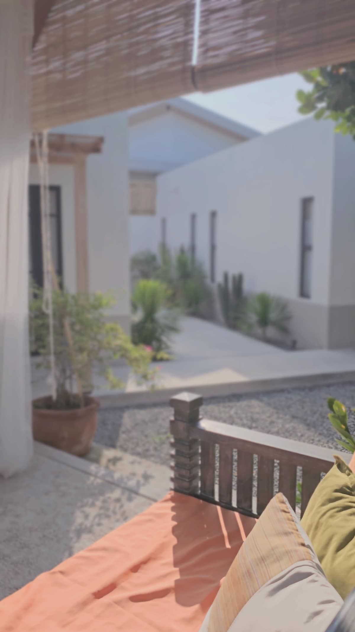 A view from a sitting area showcases a serene courtyard surrounded by greenery and stone pathways. The scene features a wooden bench with cushions and plants in pots, creating a peaceful atmosphere for relaxation.