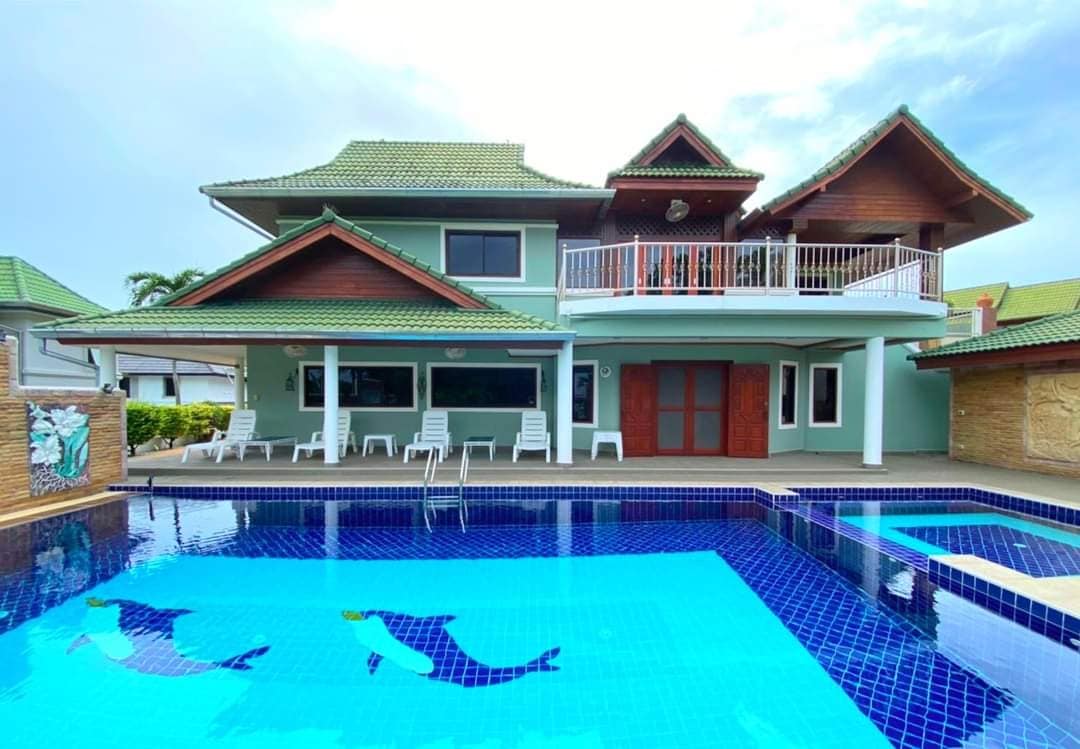 The villa features a modern architectural design with multiple gabled roofs and large windows. A spacious pool with dolphin motifs is surrounded by lounge chairs. An upper terrace extends from the building, offering outdoor relaxation space with views over the pool.