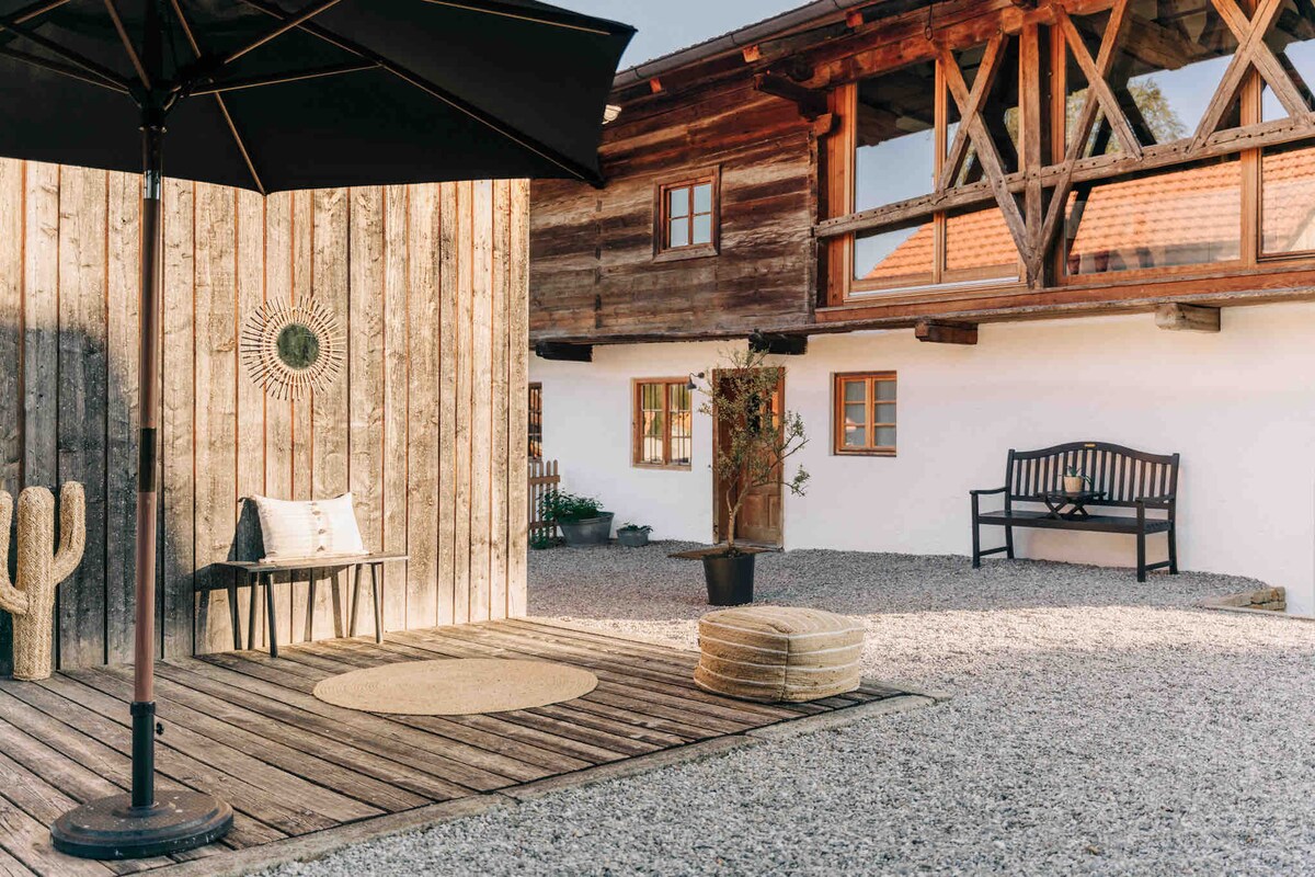 An outdoor lounge area is presented, featuring a wooden deck with a round rug and a small umbrella. There is a comfortable bench against the backdrop of a rustic farmhouse, surrounded by a gravel surface and potted plants, creating a relaxing space for guests.