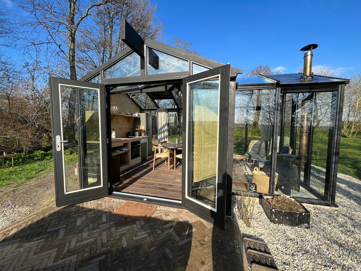 A glass living kitchen is featured, with doors open to reveal a bright and airy space. Inside, a wooden dining table and kitchenette create an inviting atmosphere. The surrounding greenery is visible, enhancing the connection to nature.