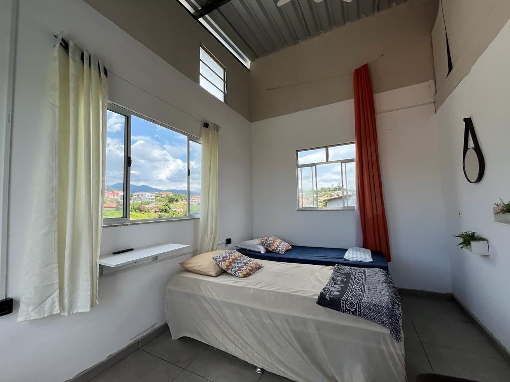 A well-lit sleeping area features two beds arranged side by side, one with a light-colored cover and decorative pillows. Large windows allow natural light to fill the space, complemented by light curtains and a vibrant orange drape. Greenery is displayed on the wall shelf.