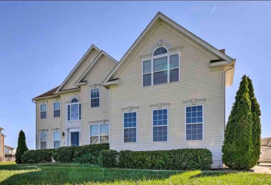 The exterior of the house features a light-colored façade with large windows. A neatly trimmed hedgerow lines the front, accompanied by two tall, conical shrubs. The structure includes a pitched roof and an arched window above the entrance, contributing to its welcoming appearance.