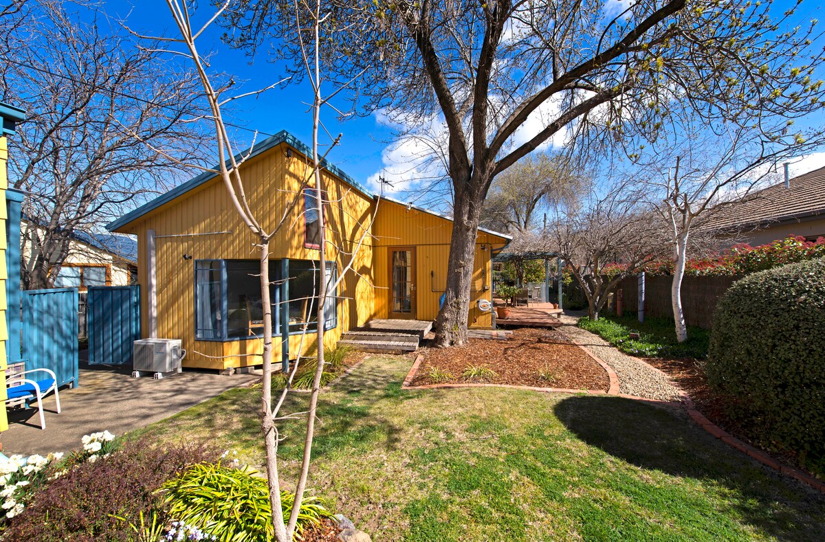 A vibrant yellow house is set against a clear blue sky, surrounded by a well-maintained garden featuring various trees and shrubs. An inviting outdoor area includes a patio with seating under the shade of a large tree. The landscape features pathways and seasonal flowers.