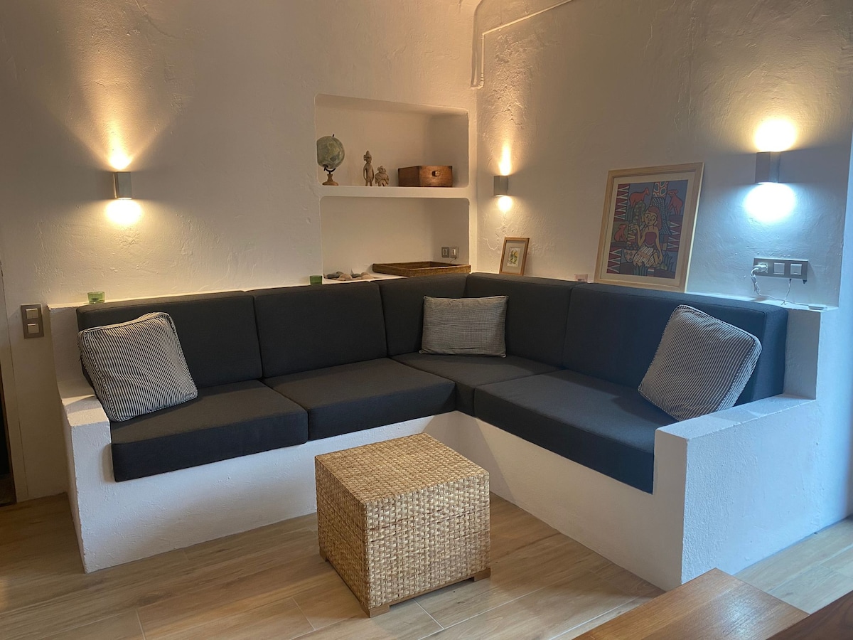 A cozy seating area is presented with a U-shaped sectional couch adorned with decorative cushions. A natural woven coffee table is centered in front of the couch, with soft wall lights illuminating the space, enhancing the modern design.