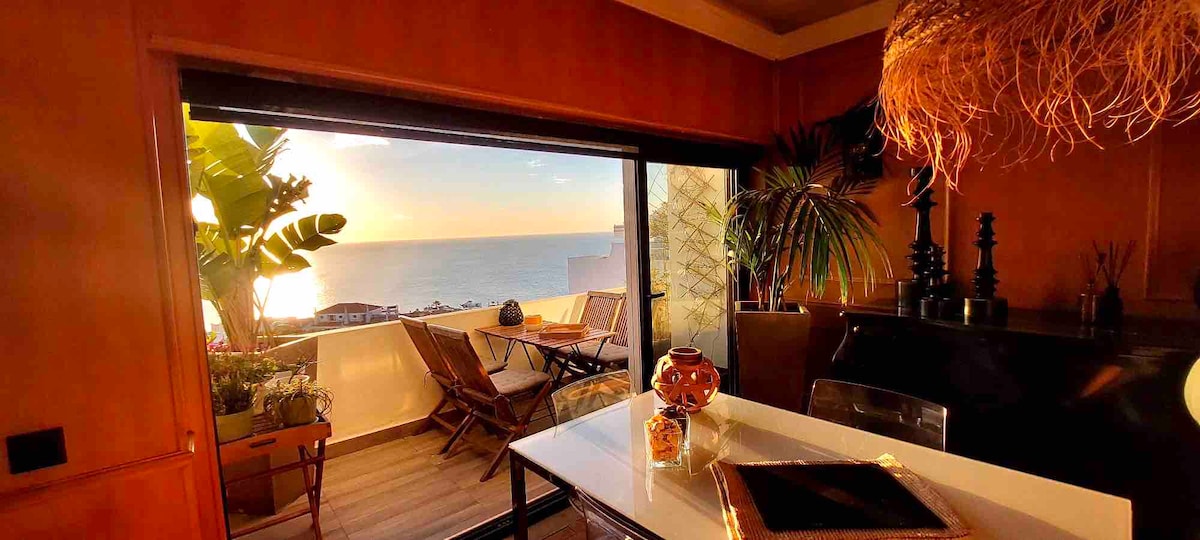A dining area is highlighted by a large window that opens to a scenic view of the ocean at sunset. A cozy table is set for meals, surrounded by wooden chairs. Decorative plants and a warm glow enhance the inviting ambiance.