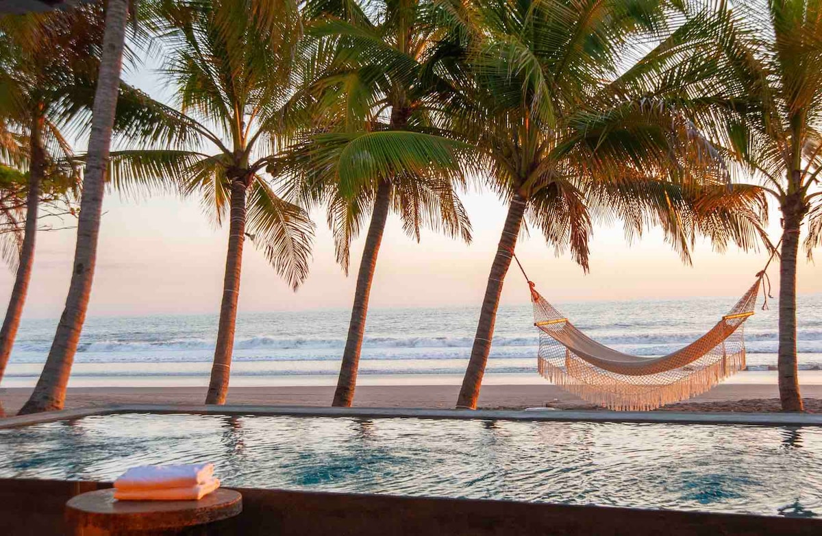 A serene swimming pool is framed by lush palm trees, overlooking a tranquil beach. A hammock sways gently between two trees, creating a relaxing atmosphere. The calm ocean waves are visible in the background, complemented by soft hues of dawn or dusk in the sky.