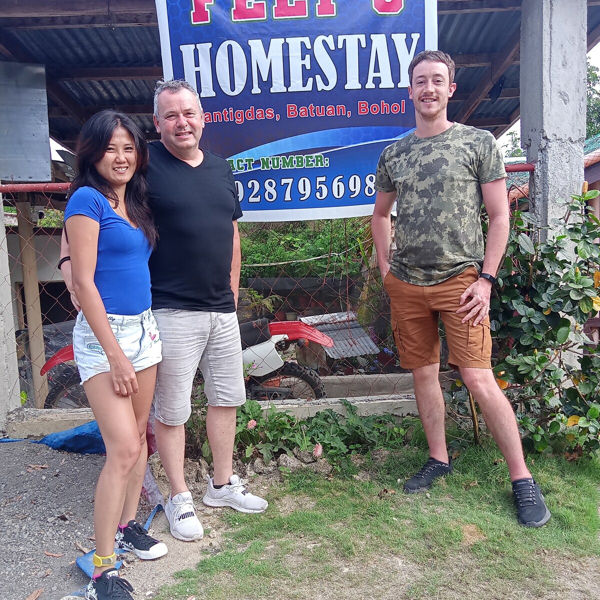 A sign for 'Peej's Homestay' is prominently displayed, indicating contact information and location. Two individuals stand nearby, casually dressed, amidst a backdrop that includes greenery. A motorbike is visible in the background, suggesting accessibility to transportation.