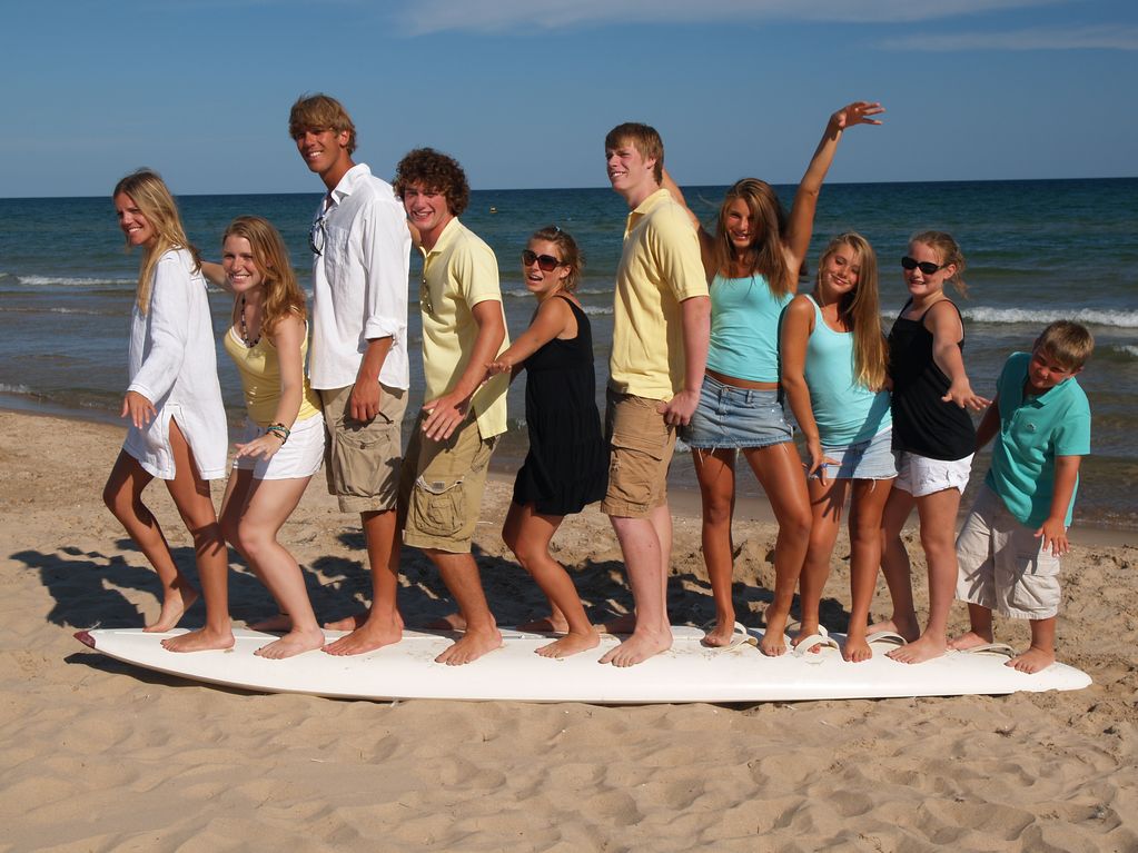 A group of individuals is standing on a surfboard on a sandy beach, with the ocean and clear blue sky in the background. Everyone is barefoot, wearing casual summer clothing. The sandy beach appears clean and inviting, creating a lively atmosphere.