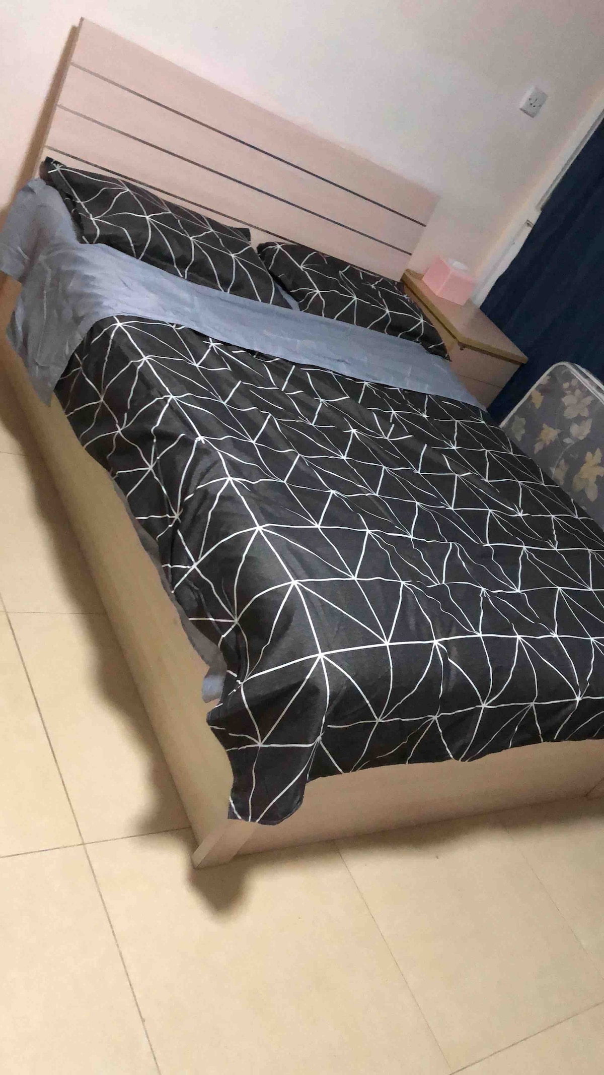 A spacious bed is centered in the room, dressed in a geometric patterned duvet in black and white. Soft grey throws are casually arranged at the foot of the bed. The walls are adorned with a light neutral tone, complemented by dark curtains on one side.