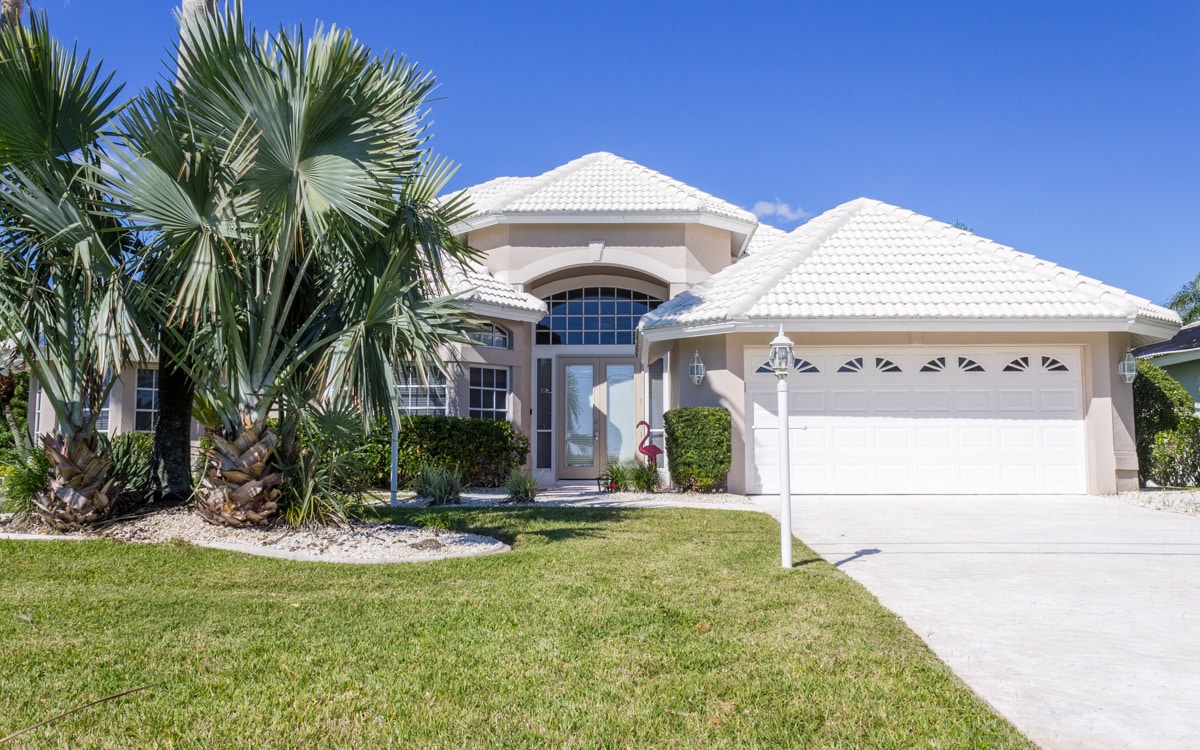 A large, elegant home features a prominent entrance with tall windows and a decorative front door. Lush palm trees and neatly trimmed landscaping frame the pathway. A spacious driveway leads to a garage with a white door, complemented by a clear blue sky overhead.