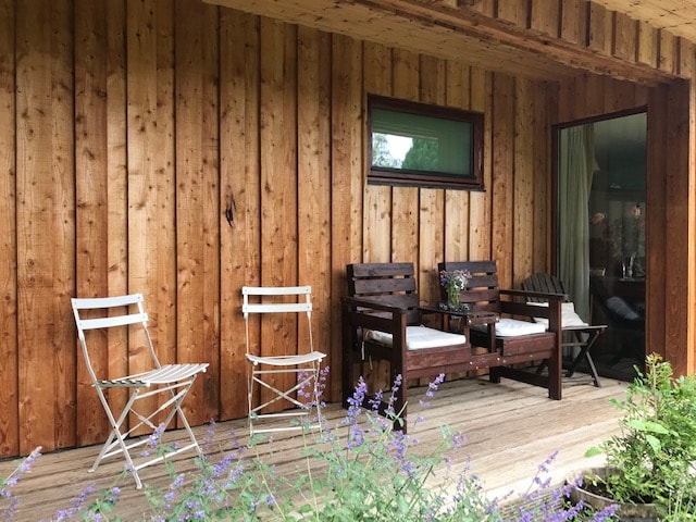 A wooden deck area features a mix of seating options, including two brown lounge chairs and a small white folding chair. Lavender plants are visible in front, enhancing the natural setting. Glass doors lead into the spacious interior, framed by warm wooden siding.