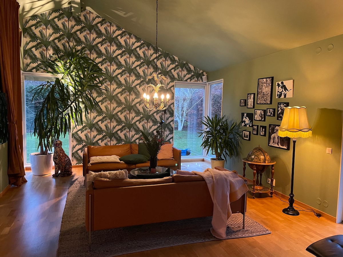 A cozy living space features a distinctive patterned wall and large windows offering views of the garden. A modern sofa is accompanied by a circular coffee table and a decorative lamp, enhancing the room's inviting appeal. Indoor plants add a touch of greenery to the atmosphere.
