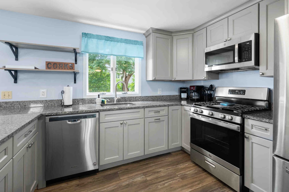 The kitchen area features modern cabinets and granite countertops, complemented by stainless-steel appliances including a dishwasher, stove, and microwave. A window provides natural light, framed by a light blue curtain. Open shelving above the countertops offers decorative storage with a 'CORKS' sign.