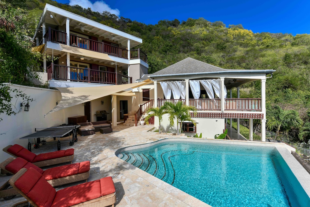 A luxurious outdoor area features a private pool surrounded by a spacious stone patio. Multiple lounge chairs are positioned nearby. The two-story house showcases large balconies and a shaded seating area, all set against a backdrop of lush greenery and hills.