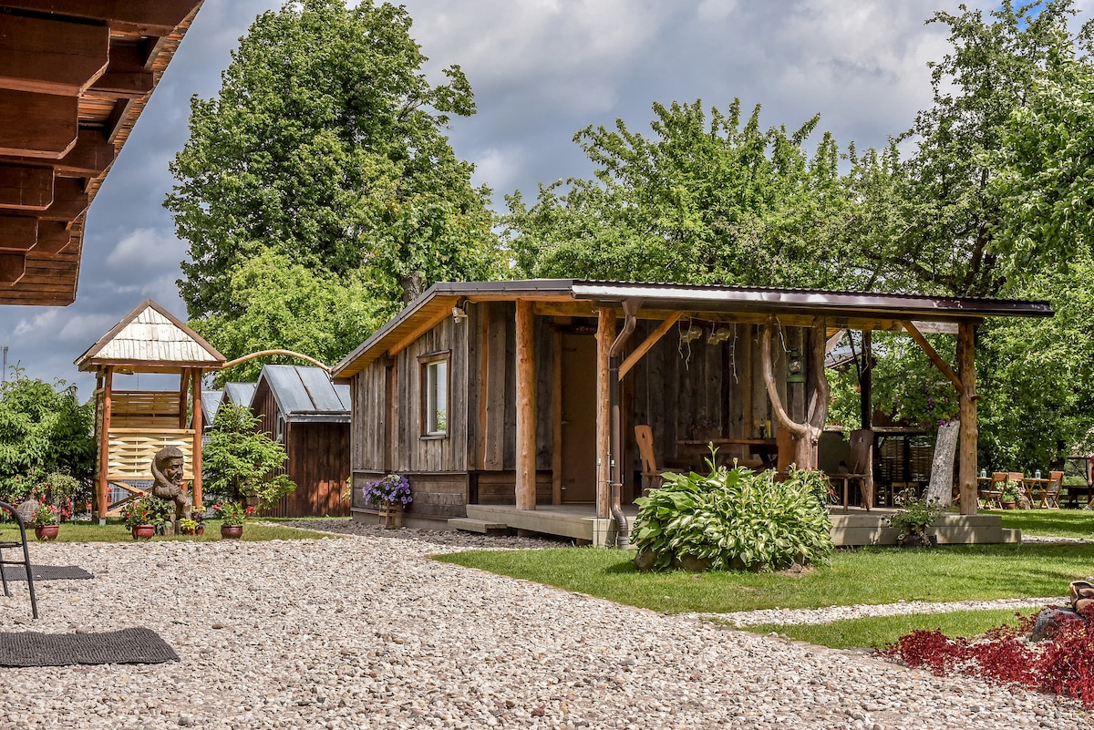 A log cabin made from Siberian wood is surrounded by lush greenery and open spaces. The structure features a covered porch and is complemented by decorative plants. Nearby, a small wooden gazebo adds to the rural charm of the environment.