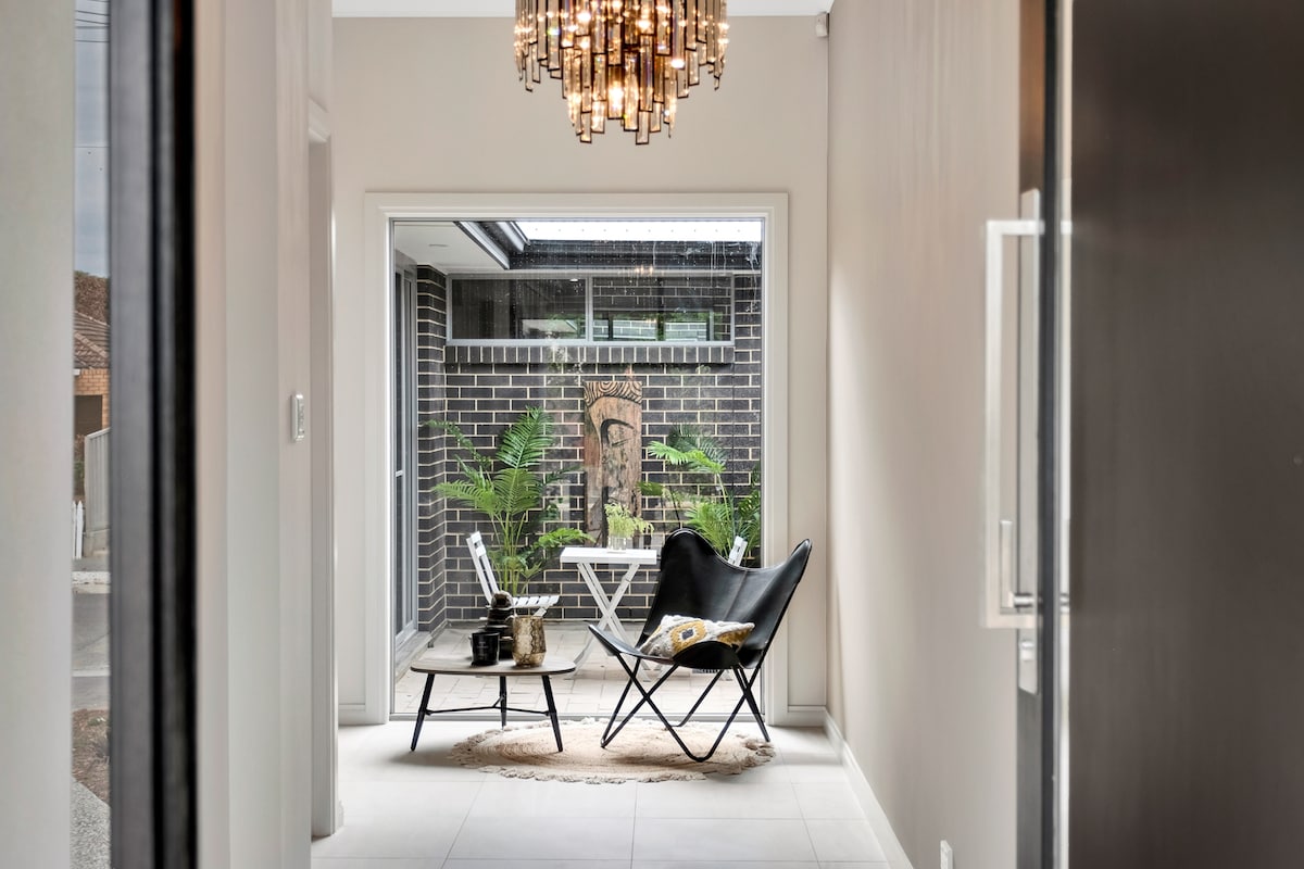 A stylish entryway is illuminated by a modern chandelier. The open space features a black lounge chair paired with a light coffee table, complemented by indoor plants visible through glass doors leading to a serene outdoor area.