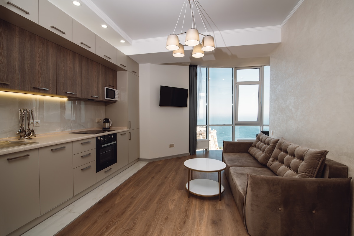 A modern kitchen area is visible, equipped with sleek cabinetry, a stove, and a microwave. A comfortable brown sofa faces a circular coffee table, and a wall-mounted television is present. Large windows allow ample natural light and showcase a view of the sea.