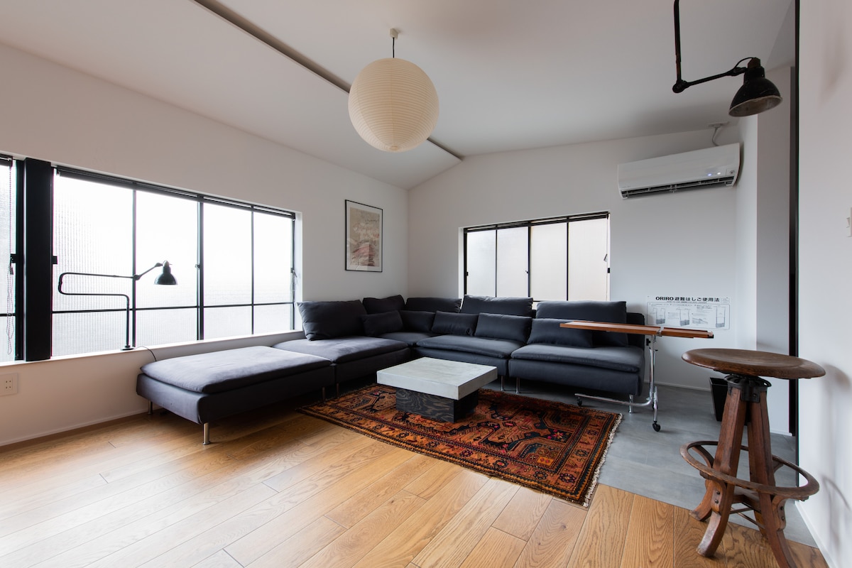 A bright living area showcases a spacious sectional sofa in deep gray, positioned around a minimalist coffee table. Two large windows allow natural light to fill the room, complemented by warm wooden flooring and an area rug, creating a cozy yet modern atmosphere.