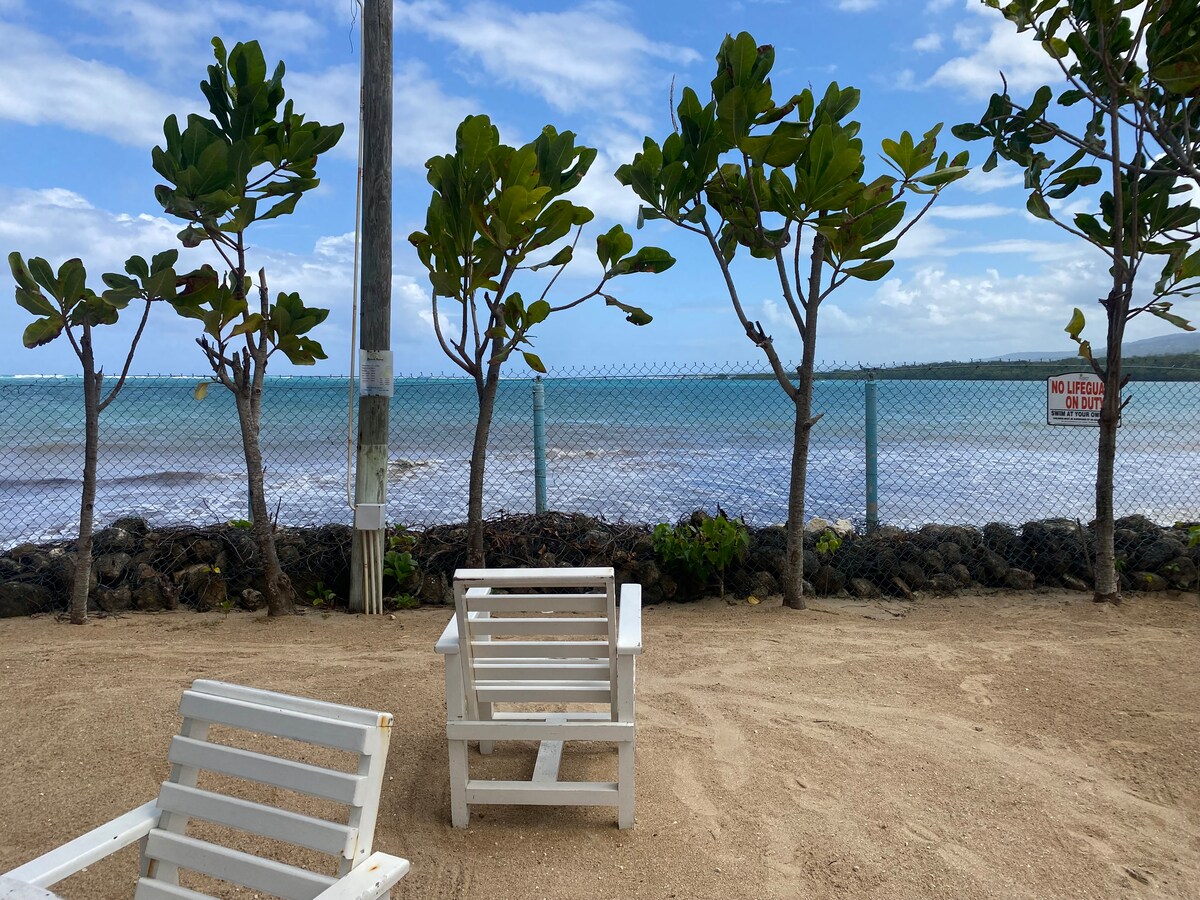 A sandy area features two white lounge chairs positioned near a low rock wall. Lush green trees frame the view of the water beyond, where gentle waves can be seen meeting the shoreline. A safety sign is visible along the fence.