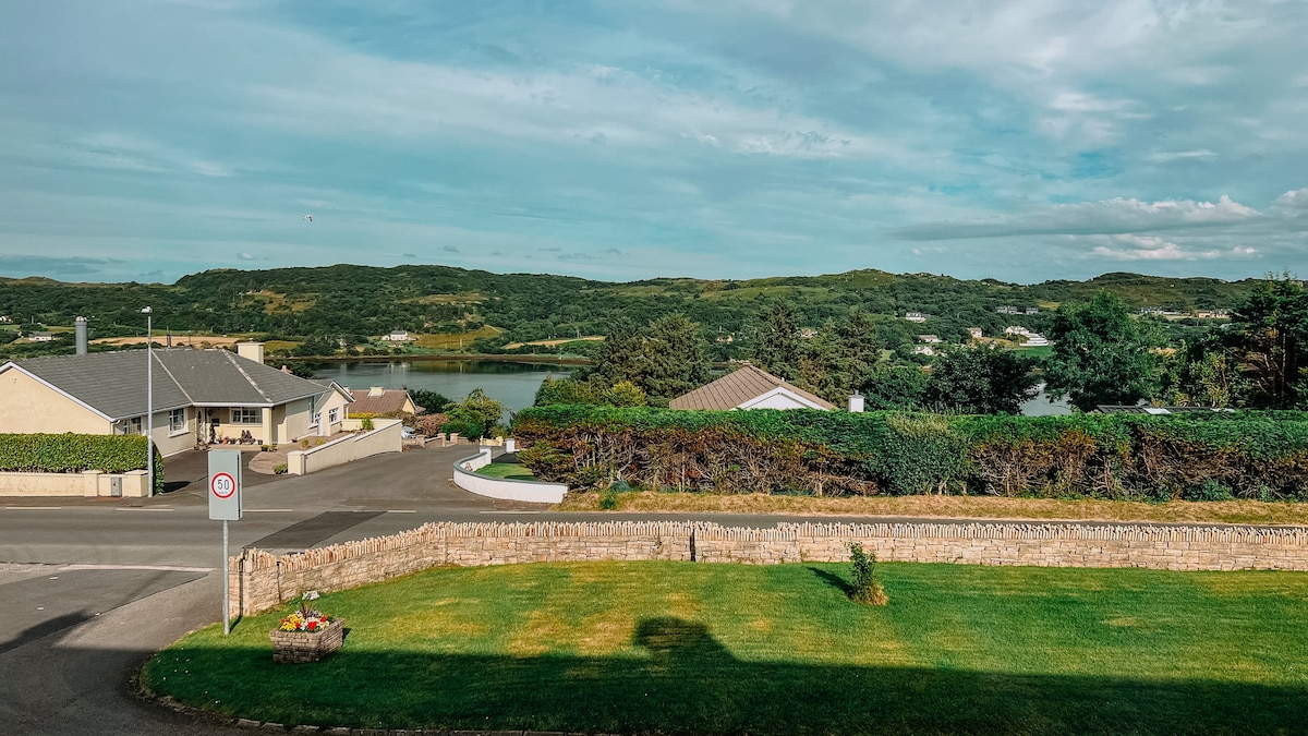 A view of the surrounding landscape features a grassy area in the foreground, with a stone wall defining the property boundary. The bay is visible in the background, along with houses nestled among lush greenery and rolling hills under a partly cloudy sky.