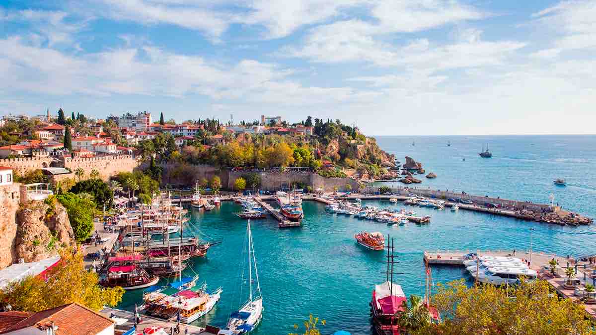 A scenic view of a coastal harbor is presented, showcasing a vibrant array of boats docked along the turquoise waters. The shore features lush greenery and colorful buildings, while the skyline reveals gentle hills and a serene blue sky dotted with clouds.